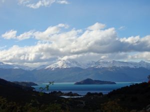 Blick über einen tiefblauen See auf die schneebedeckten Gipfel der Anden unter einem markanten Wolkenhimmel in Patagonien, Chile.