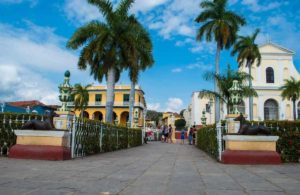 Blick auf den zentralen Plaza Mayor in Trinidad, Kuba, mit seinen historischen gelben Kolonialbauten, hohen Palmen und einer weißen Kirche unter blauem Himmel.