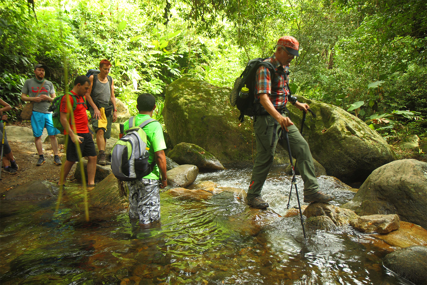 Gruppe von Wandernden watet und balanciert über einen flachen Fluss mit großen Felsblöcken im dichten Regenwald auf dem Trek zur Ciudad Perdida in Kolumbien.