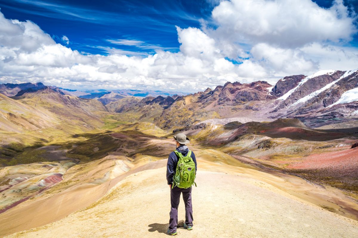 Ein Wanderer mit Rucksack blickt von einem Gipfel auf die farbenprächtige Gebirgslandschaft der Rainbow Mountains während einer Peru Rundreise.