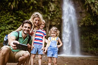 Eine vier-köpfige Familie steht vor einem Wasserfall in einem Urwald in Lateinamerika und nimmt mit dem Handy ein Selfie auf