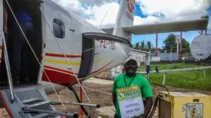 Auf dem Bild sieht man ein Kleinflugzeug. Davor steht ein Mann in grünem T-Shirt mit einem Schild, der auf eine Reisegruppe wartet.