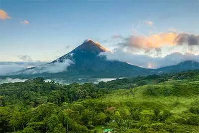 Blick auf den Vulkan Arenal, welcher von ein paar Wolken umgeben ist. Vorne sieht man den Regenwald.