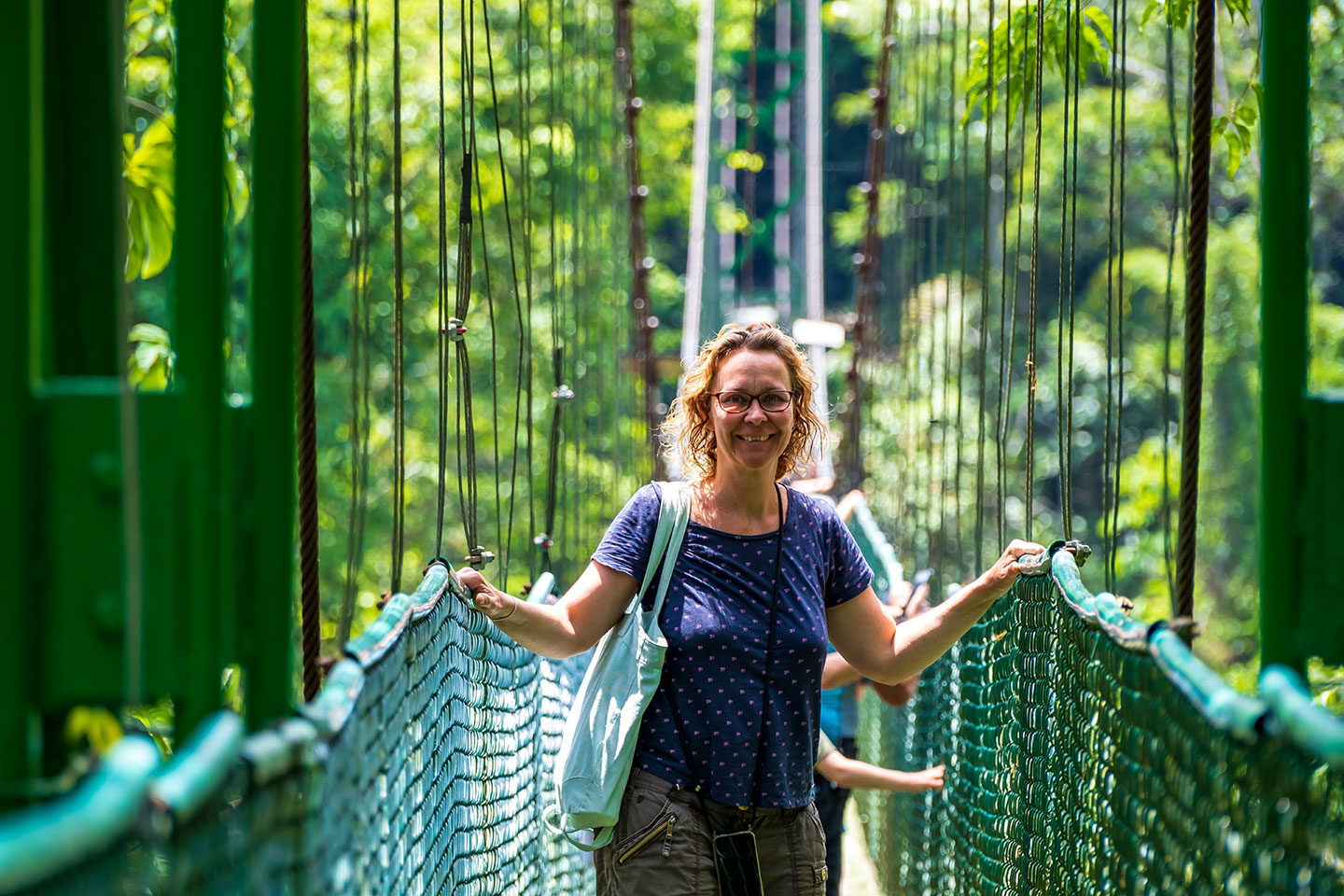 Frau überquert eine Hängebrücke im dichten Regenwald und hält sich an den seitlichen Netzen fest. Aufnahme aus einer Naturreise mit Napur Tours, die das Erlebnis auf einer Canopy-Brücke im Regenwald zeigt.