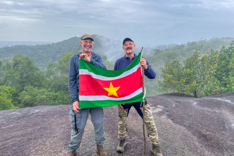Das Titelbild für den Erfahrungsbericht füber Suriname: Uwe Zündorf und sein Freund stehen auf dem Fredberg und halten eine Fahne von Suriname in den Händen. Im Hintergrund Regenwald mit Nebelschwaden.
