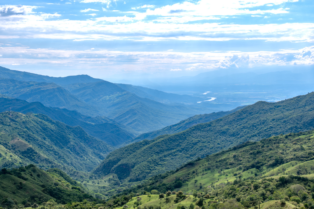 Weite grüne Berglandschaft der kolumbianischen Anden mit bewaldeten Hängen, tiefem Tal und einem Fluss in der Ferne, aufgenommen während einer Rundreise durch Kolumbien.