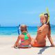 Mutter sitzt mit ihrem Kind am Strand, im Hintergrund türkisfarbenes Meer. Die Mutter lächelt in die Kamera.